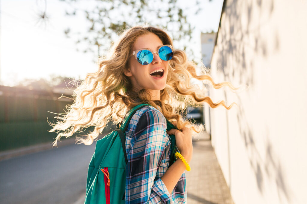 cool stylish smiling happy blond woman walking in street with backpack, curly hair, attractive, sunny, summer fashion trend, shirt, traveler, sunglasses, cheerful, backlight, positive, laughing